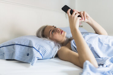Ernste junge Frau, die im Bett liegt und ihr Handy überprüft - GIOF03476