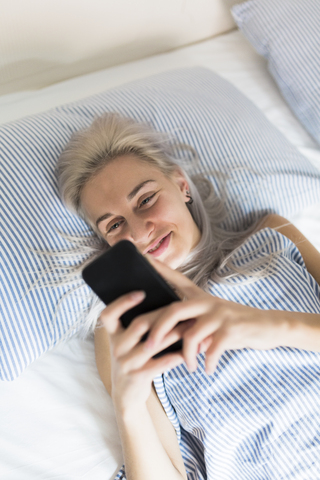 Lächelnde junge Frau, die im Bett liegt und ihr Mobiltelefon überprüft, lizenzfreies Stockfoto