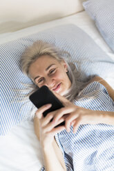 Lächelnde junge Frau, die im Bett liegt und ihr Mobiltelefon überprüft - GIOF03477
