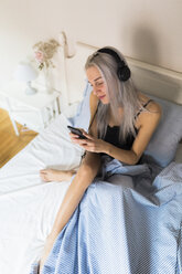 Junge Frau im Bett mit Handy und Kopfhörern - GIOF03485