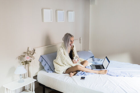 Glückliche junge Frau im Bett mit einer Tasse Kaffee und einem Laptop - GIOF03493