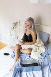 Nachdenkliche junge Frau im Bett mit einer Tasse Kaffee und einem Laptop - GIOF03494