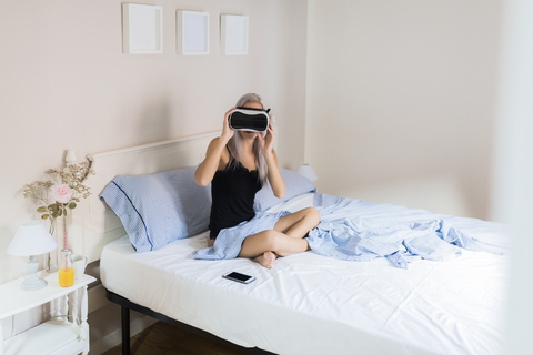 Junge Frau sitzt im Bett und trägt eine VR-Brille, lizenzfreies Stockfoto