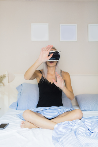 Junge Frau sitzt im Bett und trägt eine VR-Brille, lizenzfreies Stockfoto