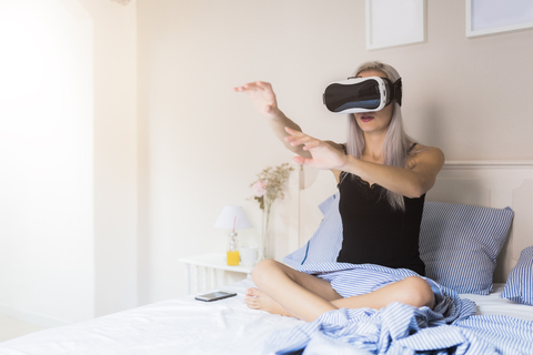 Junge Frau sitzt im Bett und trägt eine VR-Brille, lizenzfreies Stockfoto