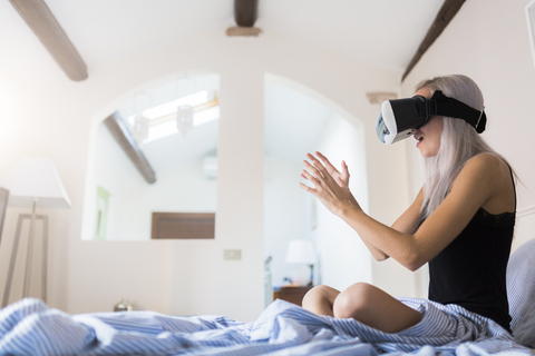 Erstaunte junge Frau sitzt im Bett und trägt eine VR-Brille, lizenzfreies Stockfoto