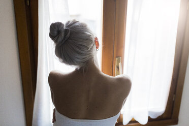 Rückansicht einer jungen Frau mit Haarknoten, die aus dem Fenster schaut - GIOF03518