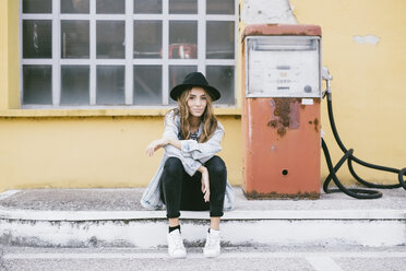 Portrait of fashionable young woman wearing hat sitting beside an old petrol pump - GIOF03533