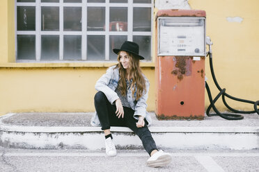 Fashionable young woman wearing hat sitting beside an old petrol pump - GIOF03534