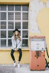 Fashionable young woman wearing hat sitting on window sill beside an old petrol pump - GIOF03535