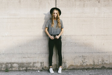 Fashionable young woman wearing hat leaning against concrete wall - GIOF03540