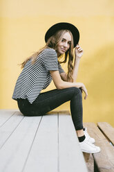 Portrait of fashionable young woman wearing hat sitting in front of yellow wall - GIOF03543