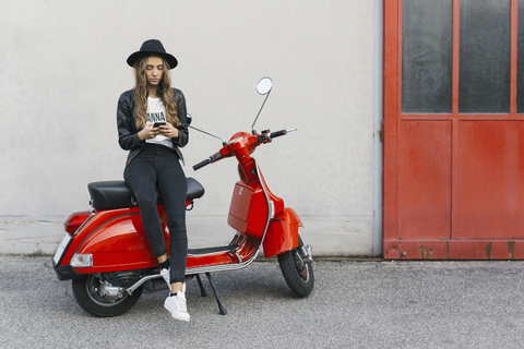 Modische junge Frau, die auf einem roten Motorroller sitzt und ein Mobiltelefon benutzt, lizenzfreies Stockfoto