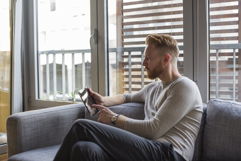 Mann benutzt Mini-Tablet auf der Couch in seinem Wohnzimmer, lizenzfreies Stockfoto