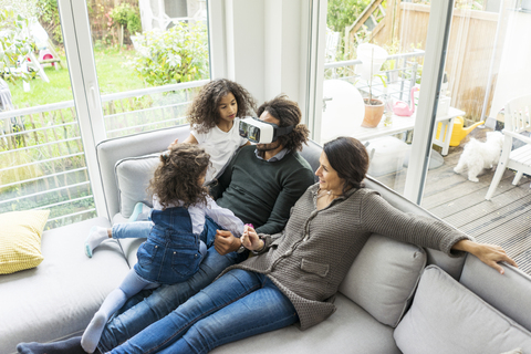 Glückliche Familie sitzt auf der Couch, der Vater trägt eine VR-Brille, lizenzfreies Stockfoto