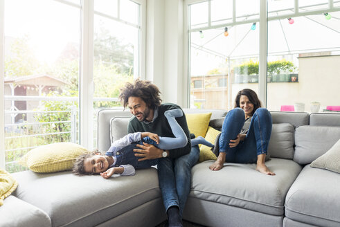 Happy family sitting on couch, father tickling his laughing daughter - MOEF00325