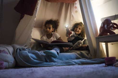 Zwei Schwestern sitzen in einem dunklen Kinderzimmer, spielen mit einer Taschenlampe und lesen ein Buch - MOEF00335