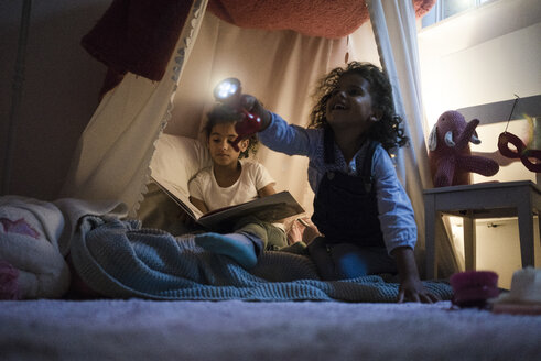 Zwei Schwestern sitzen in einem dunklen Kinderzimmer, spielen mit einer Taschenlampe und lesen ein Buch - MOEF00336