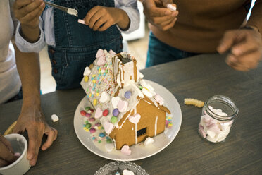 Family pasting gingerbread house in kitchen for Christmas with sweets - MOEF00379