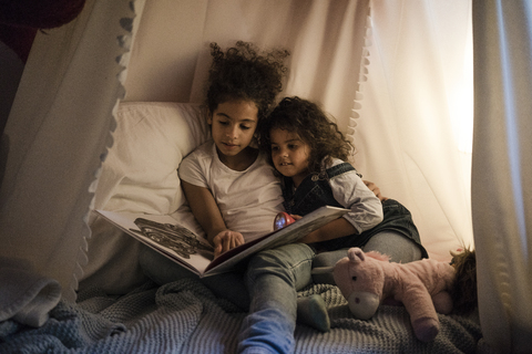 Zwei Schwestern sitzen in einem dunklen Kinderzimmer und lesen ein Buch, lizenzfreies Stockfoto