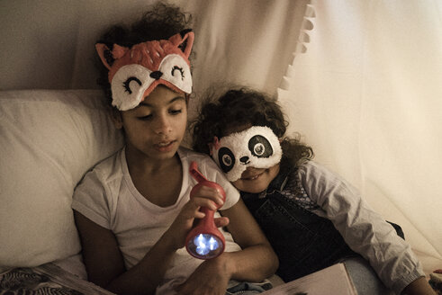 Zwei Schwestern sitzen in einem dunklen Kinderzimmer, spielen mit einer Taschenlampe und lesen ein Buch - MOEF00406