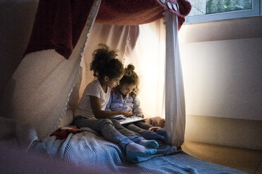 Zwei Schwestern sitzen in einem dunklen Kinderzimmer und schauen auf ein digitales Tablet - MOEF00410