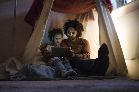 Vater und Tochter sitzen in einem dunklen Kinderzimmer und schauen auf ein digitales Tablet, lizenzfreies Stockfoto