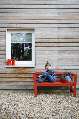 Mädchen sitzt auf einer roten Bank vor einer Holzfassade, lizenzfreies Stockfoto