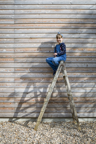 Lachendes Mädchen, das auf einer Leiter vor einer Holzfassade sitzt, lizenzfreies Stockfoto