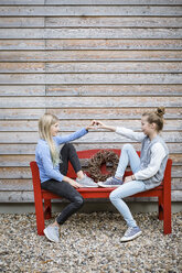 Two best friends sitting on a red bench in front of a wooden facade forming heart with their hands - OJF00216