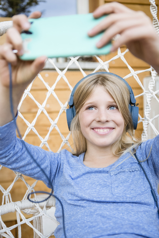 Porträt eines lachenden Mädchens mit Kopfhörern, das ein Selfie mit einem Smartphone in einem Hängesessel macht, lizenzfreies Stockfoto