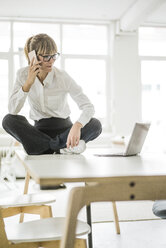 Businesswoman sitting on desk in office with laptop and cell phone - JOSF01945