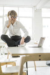 Geschäftsfrau sitzt auf dem Schreibtisch im Büro mit Laptop und Mobiltelefon - JOSF01945