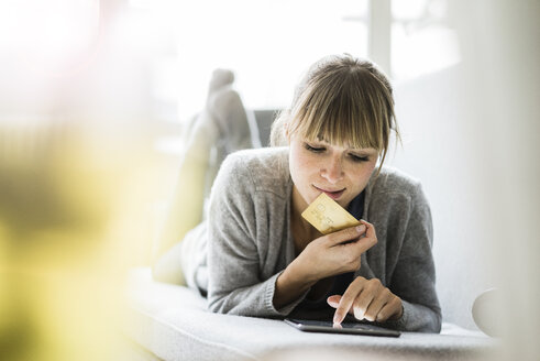 Woman lying on couch with credit card and tablet - JOSF01948