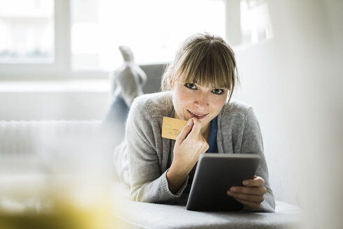 Smiling woman lying on couch with credit card and tablet - JOSF01955