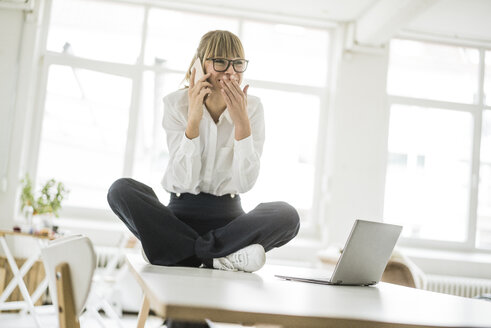 Happy businesswoman sitting on desk in office talking on cell phone - JOSF01972