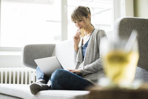 Smiling woman sitting on couch with credit card and laptop - JOSF01996