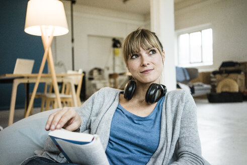 Smiling woman sitting in beanbag with book and headphones - JOSF02004