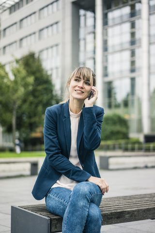 Lächelnde Geschäftsfrau, die auf einer Bank sitzt und mit einem Handy im Freien telefoniert, lizenzfreies Stockfoto