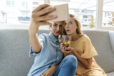 Zwei junge Frauen sitzen auf der Couch und machen ein Selfie mit Süßigkeiten - KNSF03040