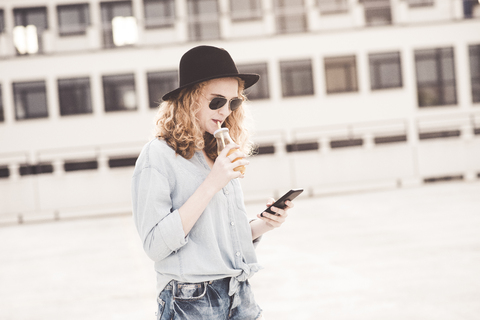 Junge Frau trinkt ein Getränk und benutzt dabei ein Handy, lizenzfreies Stockfoto