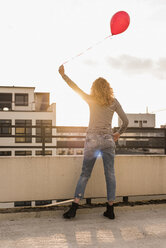 Rückenansicht eines jungen Mannes mit rotem Ballon auf der Dachterrasse bei Sonnenuntergang - UUF12344