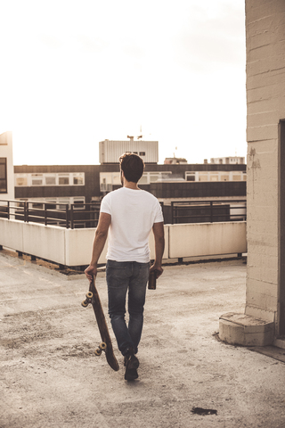 Rückenansicht eines jungen Mannes mit Skateboard und Bierflasche auf einer Dachterrasse in der Abenddämmerung, lizenzfreies Stockfoto