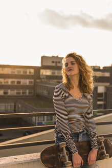 Porträt einer jungen Frau mit Skateboard, die den Sonnenuntergang auf einer Dachterrasse genießt - UUF12355