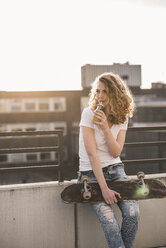 Junge Frau mit Skateboard und Getränk auf Dachterrasse bei Sonnenuntergang - UUF12359