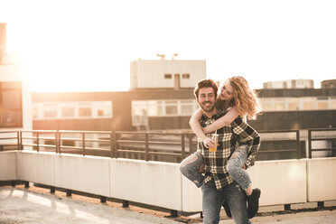 Porträt eines jungen Mannes, der seine Freundin bei Sonnenuntergang auf der Dachterrasse huckepack nimmt - UUF12361