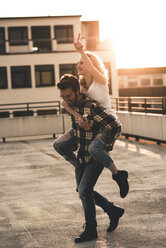 Glücklicher junger Mann, der seine Freundin bei Sonnenuntergang auf der Dachterrasse huckepack nimmt - UUF12363