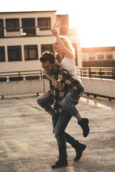 Happy young man giving his girlfriend a piggyback ride on roof terrace at sunset - UUF12363