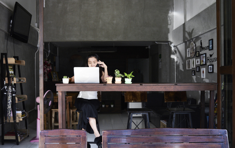 Woman sitting at wooden table using laptop and cell phone stock photo