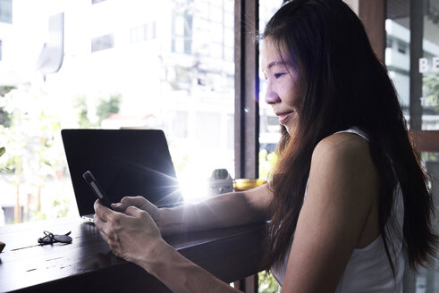 Smiling woman with laptop at the window using cell phone - IGGF00206